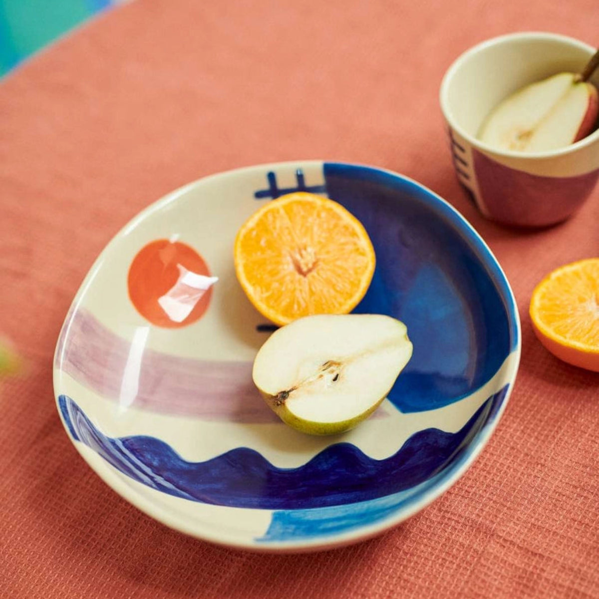 A halved pear and a halved orange rest in the Sun + Sea Serving Bowl by Donna Wilson, its colorful, hand-painted abstract patterns popping against a textured coral tablecloth. In the background, a small cup with fruit adds to the playful aesthetic inspired by Donna Wilson’s signature style.