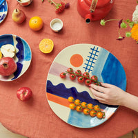 A hand arranges tomatoes on the Donna Wilson Sun + Sea Serving Platter. The table is set with a red cloth, assorted fruit, dishes featuring abstract Donna Wilson designs, a red teapot, and flowers.