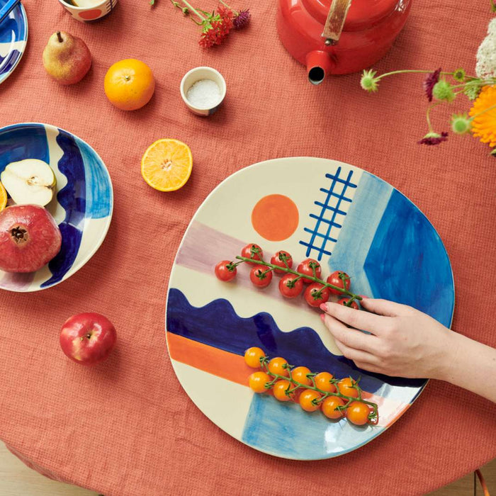 A hand arranges tomatoes on the Donna Wilson Sun + Sea Serving Platter. The table is set with a red cloth, assorted fruit, dishes featuring abstract Donna Wilson designs, a red teapot, and flowers.