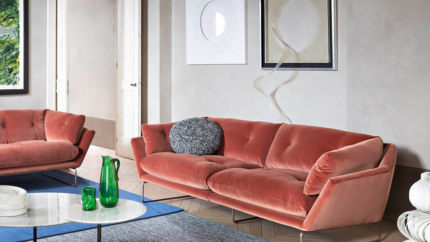 A modern living room features light gray walls, a Saba New York Suite XL 4-Seater Velvet Sofa, loveseat, blue rug, white marble coffee table with green glass vases, abstract art, and natural light streaming through the open doorway.